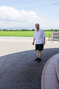 An elderly man stands on a sidewalk in front of a building. The sky is blue and there are some clouds