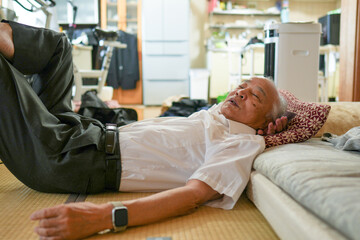 An older man is laying on a bed with a pillow. He is wearing a white shirt and gray pants