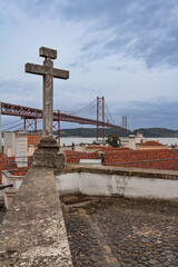 Scenic view of Lisbon's Tagus River and 25 de Abril Bridge framed by an ancient stone cross and red tiled rooftops under a cloudy sky.
