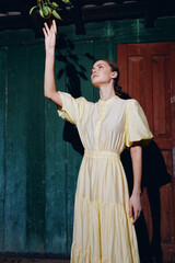 Obraz premium Woman in yellow vintage dress reaching for hanging fruit indoors authenticity inclusivity portrait with dramatic shadow and rustic wall