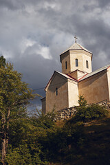Fototapeta premium Ancient stone church perched on a hillside under a dramatic cloudy sky, surrounded by trees, showcasing historic architecture, and a tranquil rural landscape with a weathered tower