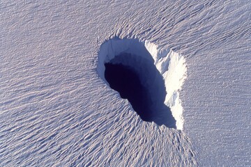 A deep circular hole in a snowy expanse reveals sharp edges and a dark center, reflecting light off the snow
