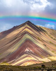 Vivid mountainous landscape of layered colors, crowned by a bright full rainbow under cloudy sky