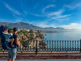 Active father carrying a toddler in a hiking backpack stands by a railing admiring scenic coastline and mountain views in Nerja, Spain, enjoying a family summer vacation on the sunny Costa del Sol.