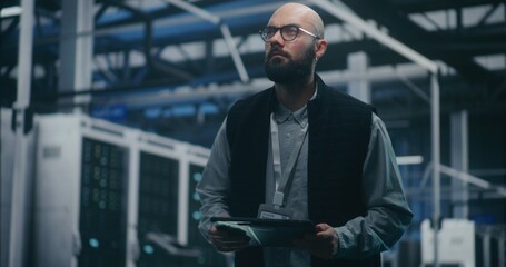 Fototapeta premium Engineer Checking System Metrics on Digital Tablet. Bearded IT Engineer in Dark Vest and Glasses Monitors System Metrics on Digital Tablet Amidst Server Racks. Cloud Auditing, and Technical Analysis.