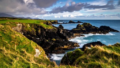 A coastal scene showing a green hillside overlooking a rocky shoreline. Water rushes over rocks, under a partly cloudy sky