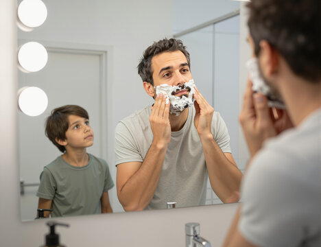 A father and his son share a special moment in the bathroom as the dad shaves his face while the boy watches curiously, showcasing the beauty of fatherhood and bonding between them
