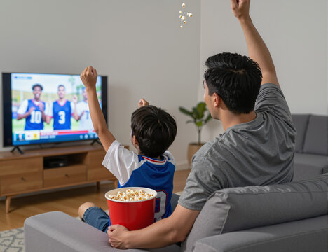 A father and son bonding while watching sports on television, celebrating a game together with excitement and joy, enjoying popcorn and sharing a memorable moment in the living room