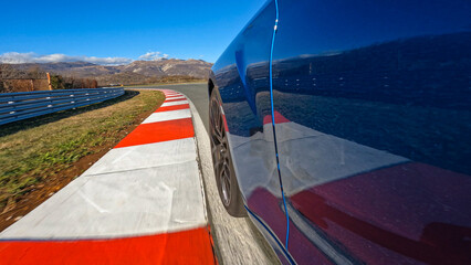 LOW ANGLE, CLOSE UP: Metallic blue sportscar speeds along the empty racetrack on an idyllic sunny day. Action shot of a brand new luxury car racing on a closed circuit on a perfect sunny afternoon. © helivideo