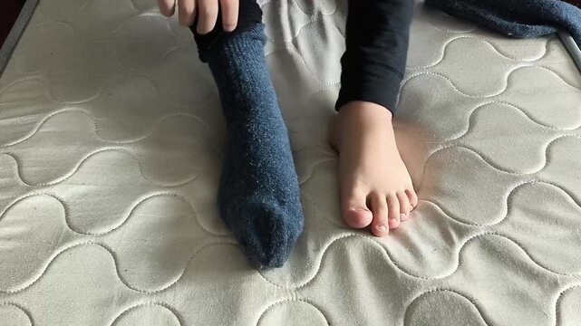 A teenage boy puts on warm wool socks while sitting on the bed. Concept of a cold floor at home