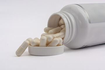 White pill bottle with white capsules spilling into the cap on white background