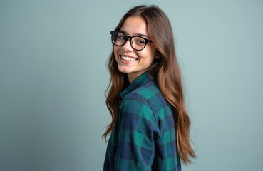 Fototapeta premium Young woman with glasses smiles happily over her shoulder. She wears a green plaid shirt against a plain grey backdrop, exuding a friendly, confident vibe. Her long hair falls loosely around her.
