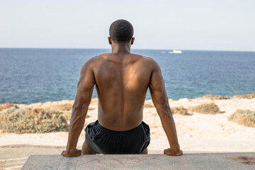 Muscular man performing bench dips by the sea. Shirtless African American athlete doing triceps dips exercise on concrete bench during outdoor workout by the ocean.