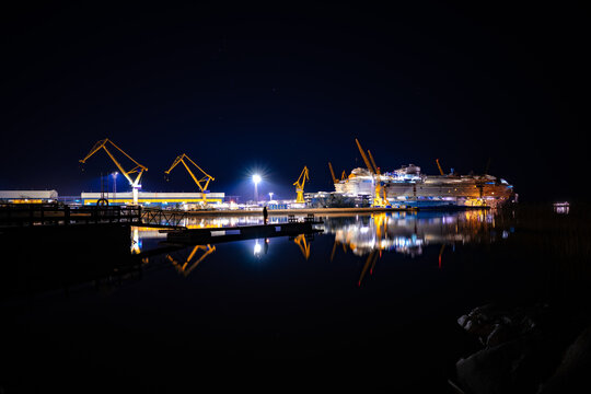 Night view of a massive cruise ship under construction in a modern shipyard with industrial cranes and water reflections.