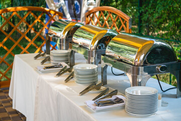 Buffet catering service setting up chafing dishes, stacked plates, and serving utensils on a white tablecloth, prepared for an outdoor event like a garden party or celebration