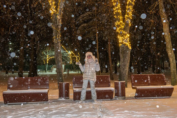 Mature adult female in winter clothes on the background of old town park benches. Night illumination. Snowfall.