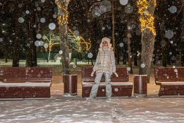 Mature adult female in winter clothes on the background of old town park benches. Night illumination. Snowfall.