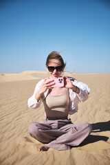 Young woman wearing sunglasses and light clothing sits cross-legged on golden sand dunes under clear blue sky and takes a picture on her smartphone. Sand dunes in Sahara desert, Siwa oasis, Egypt