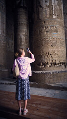 Young woman taking a picure of massive ancient stone columns covered with carved reliefs. Temple of Hathor, Dendera Temple complex, Qena, Egypt