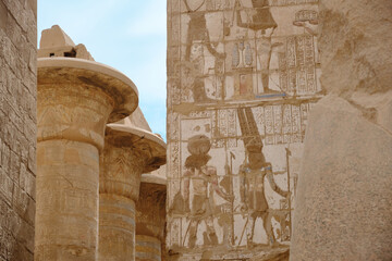 Ancient Egyptian stone reliefs carved into temple walls, depicting human figures and symbolic scenes beside massive sandstone columns. Hieroglyphic carvings on ancient monument under natural daylight