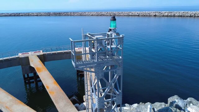 Close-up view of a galvanized metal tower housing a green navigation light at the entrance to a port facility protected by a breakwater on the calm waters of the St. Lawrence River. Matane, Quebec.
