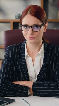 Close-up portrait of beautiful millennial businesswoman sitting at desk looking at camera in modern office. Successful young caucasian woman in glasses with red hair working at office and posing to