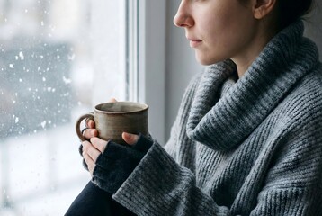 Pensive young woman in cozy sweater looking at snowfall with hot drink by the window.