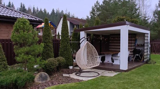 A wooden gazebo with a table, benches and a metal roof and a pine tree are decorated with glowing garlands for Christmas and New Year. There are trees, swings and houses nearby. Cloudy and snowless