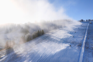 Wide view of snowmaking system with snow cannons operating on ski slope in winter resort