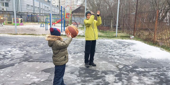 A child is throwing a basketball while a teenager watches him at a winter playground. - Powered by Adobe