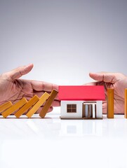 Protective Hands Shielding Toy House from Falling Blocks &ndash; Financial Security Concept