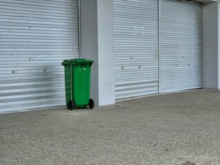 Green wheeled trash bin standing outdoors against metal wall in urban industrial setting