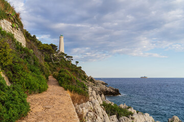 Scenic stone coastal path leading toward the historic lighthouse of Saint-Jean-Cap-Ferrat under a cloudy sky in the French Riviera.