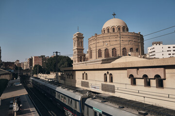 Wide view of Church of St. George rising above urban railway line, with trains passing below and city buildings in the background. Old or Coptic Cairo, Egypt