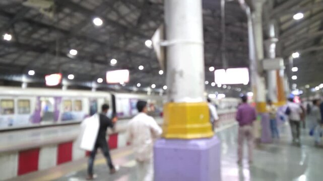 A blurred video still captures a busy Mumbai local train station platform. Commuters are actively moving about the platform as a local train appears to be departing or arriving. No logo, brand visible