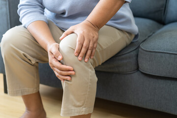 Body arthritis muscles stiff problem, aged woman suffering on leg, pain knee from disease chronic, hand massage rubbing joint injury, mature hurt or sore sitting on sofa at home. Health care people.