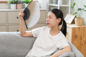 Suffering summer heat wave stroke, hot weather, tired asian young woman feeling discomfort sweaty, sitting on couch in living room, hand fan in blowing, wave fan to ventilation when high temperature.