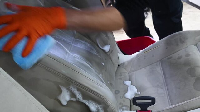 A professional vehicle detailing specialist meticulously cleans the cream-colored leather car seat with a special cleaning foam and sponge to remove stains.