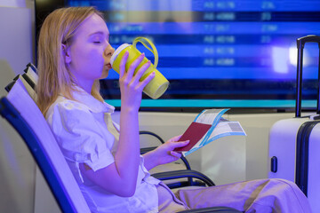 A traveler waits in an airport terminal, drinking from a mug, holding their passport and boarding passes next to their suitcase in front of the flight information board