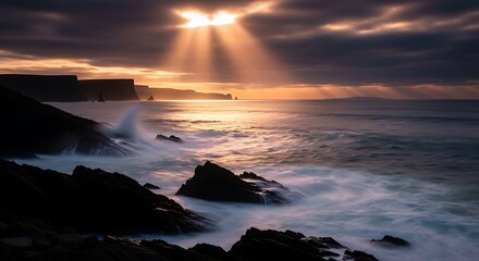 Sunbeams pierce storm clouds over a rocky ocean coast.