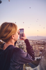 Young woman captures scenic view of colorful hot air balloons rising over dramatic rocky landscape of Cappadocia at sunrise