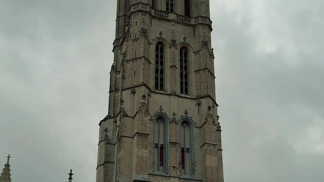 Tilt up view of the majestic Saint Bavo's Cathedral tower in Ghent, Belgium. Historic Gothic architecture rises high against a cloudy sky in the city center.

