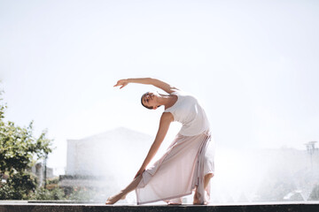 Female dancer stretching gracefully on a concrete ledge, feeling the freedom of movement and body flexibility in a serene outdoor setting with a sparkling water fountain backdrop