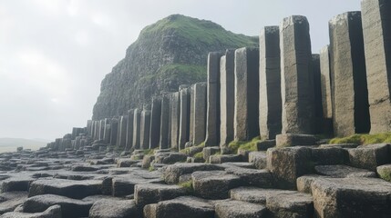 A repeating pattern of smooth grey basalt columns arranged on a rocky ground with a green grassy hill in the background under an overcast sky