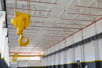 Industrial workshop with an overhead crane fire safety system red pipes. A bright yellow crane hook is suspended from the ceiling. Concept of industry, manufacturing, engineering, interior