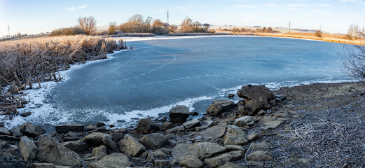 Captured in Highland, Czech Republic, a tranquil, icy lake is bordered by frost-kissed reeds and rocks, reflecting crisp winter sunlight on a peaceful morning.
