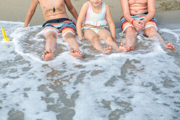 three children sitting on sandy beach with feet in shallow ocean water, enjoying gentle waves and summer fun together during family vacation, kids playing seaside with sand and water, childhood joy