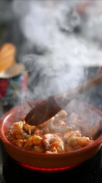 Vertical cooking of garlic shrimp sizzling in clay pot stirred with wooden spoon and rising steam , recipe for prawns in pil pil sauce