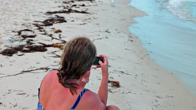 Close up view of woman photographing shoreline while sitting on sandy Miami Beach of Atlantic Ocean. USA.