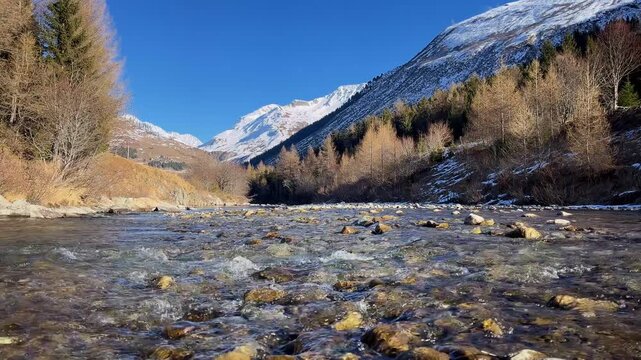 Mountain river in an alpine valley. Clear stream with stones, snow-covered peaks, and forested slopes. Winter scenery,  Outdoor travel background. Reuss River, Andermatt region, Switzerland.